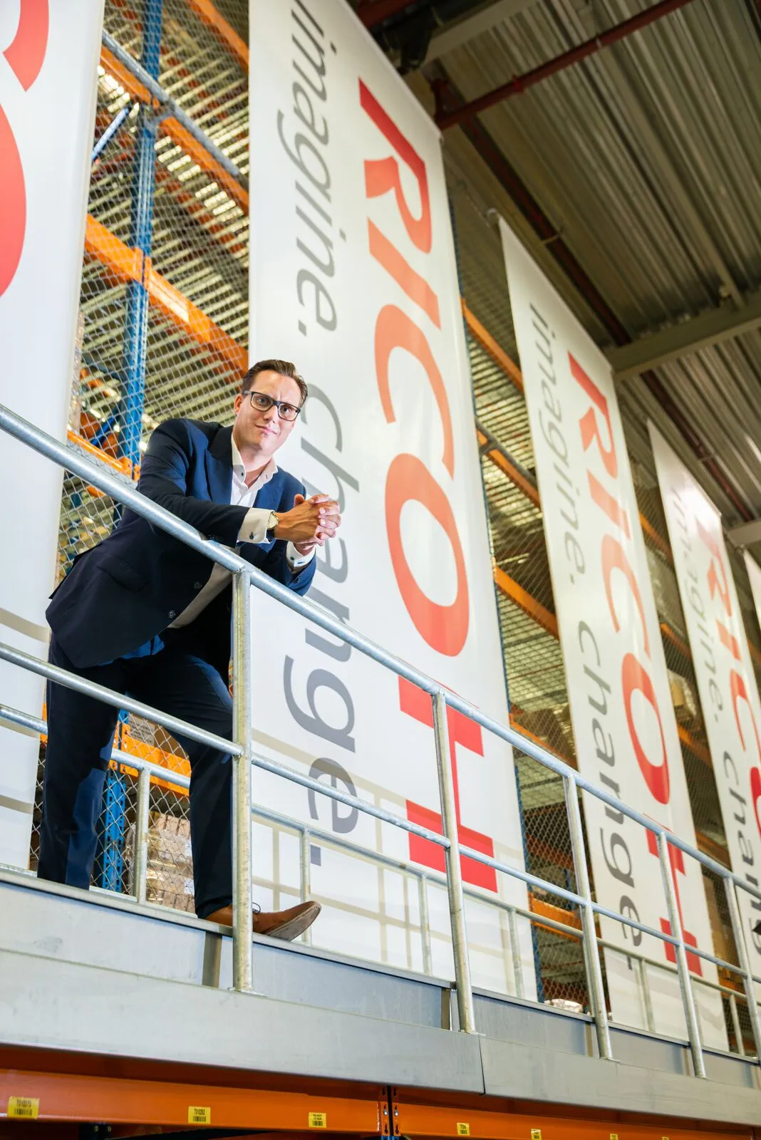 Martijn Spee leaning on a mezzanine railing in front of Ricoh banners in a warehouse