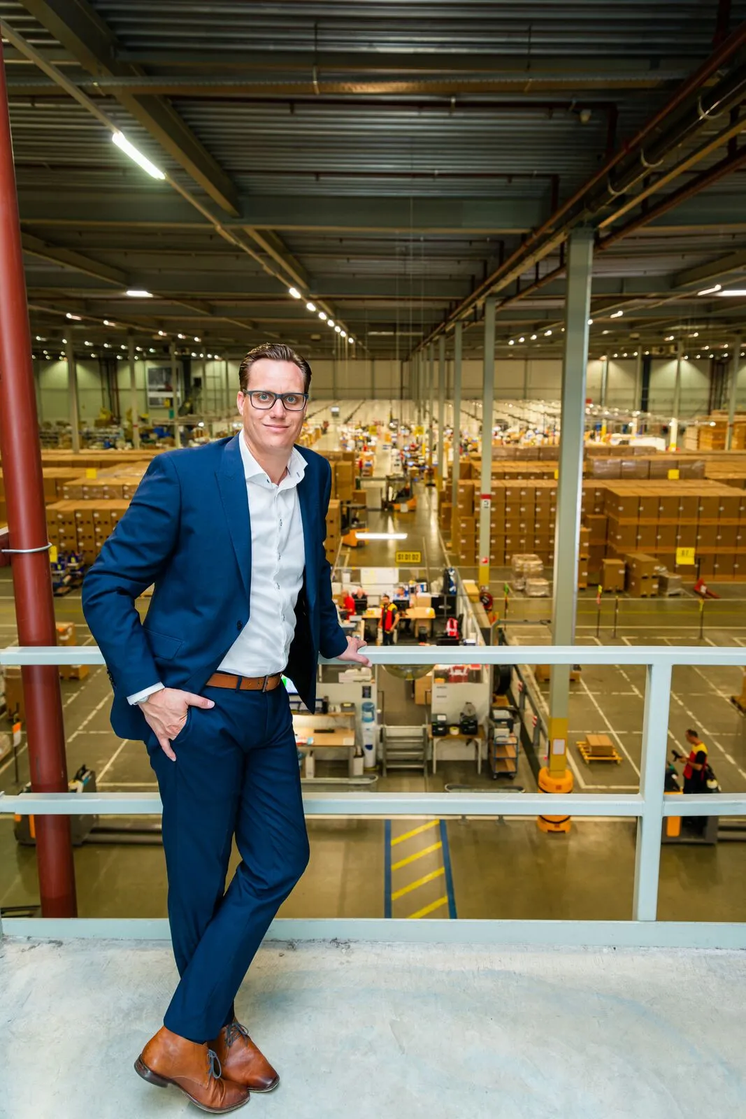 Martijn Spee standing at a mezzanine railing overlooking a large warehouse floor of stacked boxes